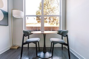 A corner room with a round table and two chairs, a floor lamp, and a window with a view of a wooden fence and trees. - Blueground | Chevy Chase, rooftop, nr Rock Creek Park (Chevy Chase)