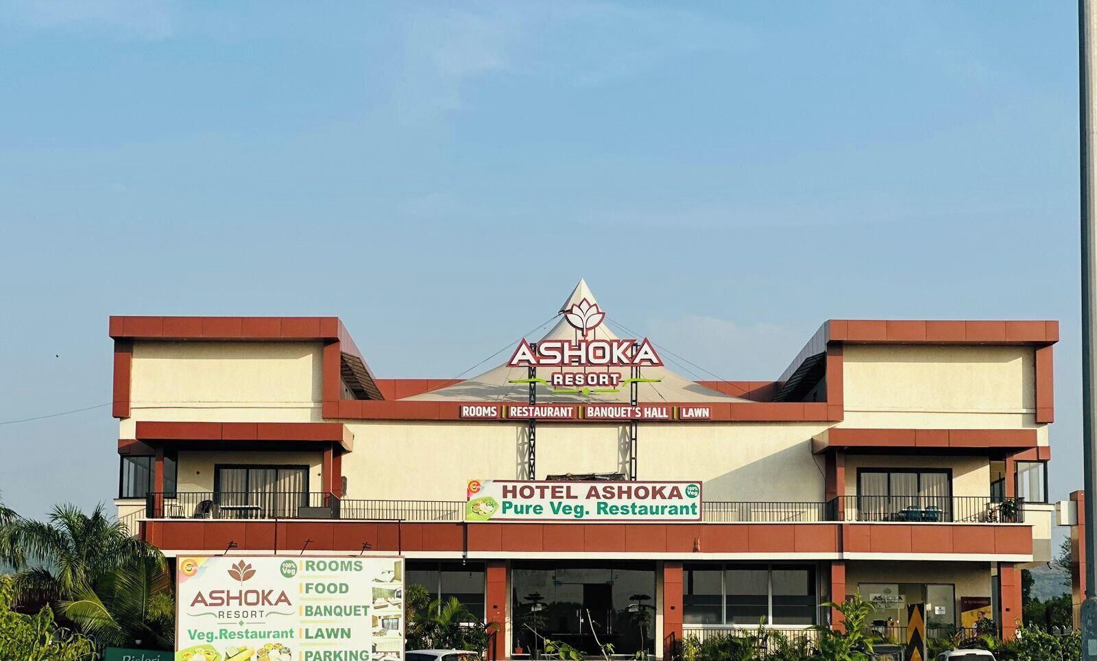 A two-story building with a sign for Hotel Ashoka Resort, featuring amenities such as rooms, a restaurant, banquet hall, and lawn.