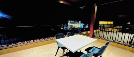 Un restaurante con una mesa blanca, sillas azules, suelo de baldosas y vistas de la ciudad de noche.