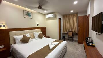 A hotel room with a bed, bedside table, lamp, air conditioning unit, desk, chair, and a television.