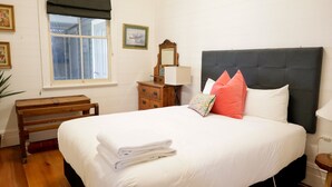 Una cama bien hecha con sábanas blancas y una toalla doblada, un banco de madera, un aparador con espejo y una ventana con cortinero oscuro.