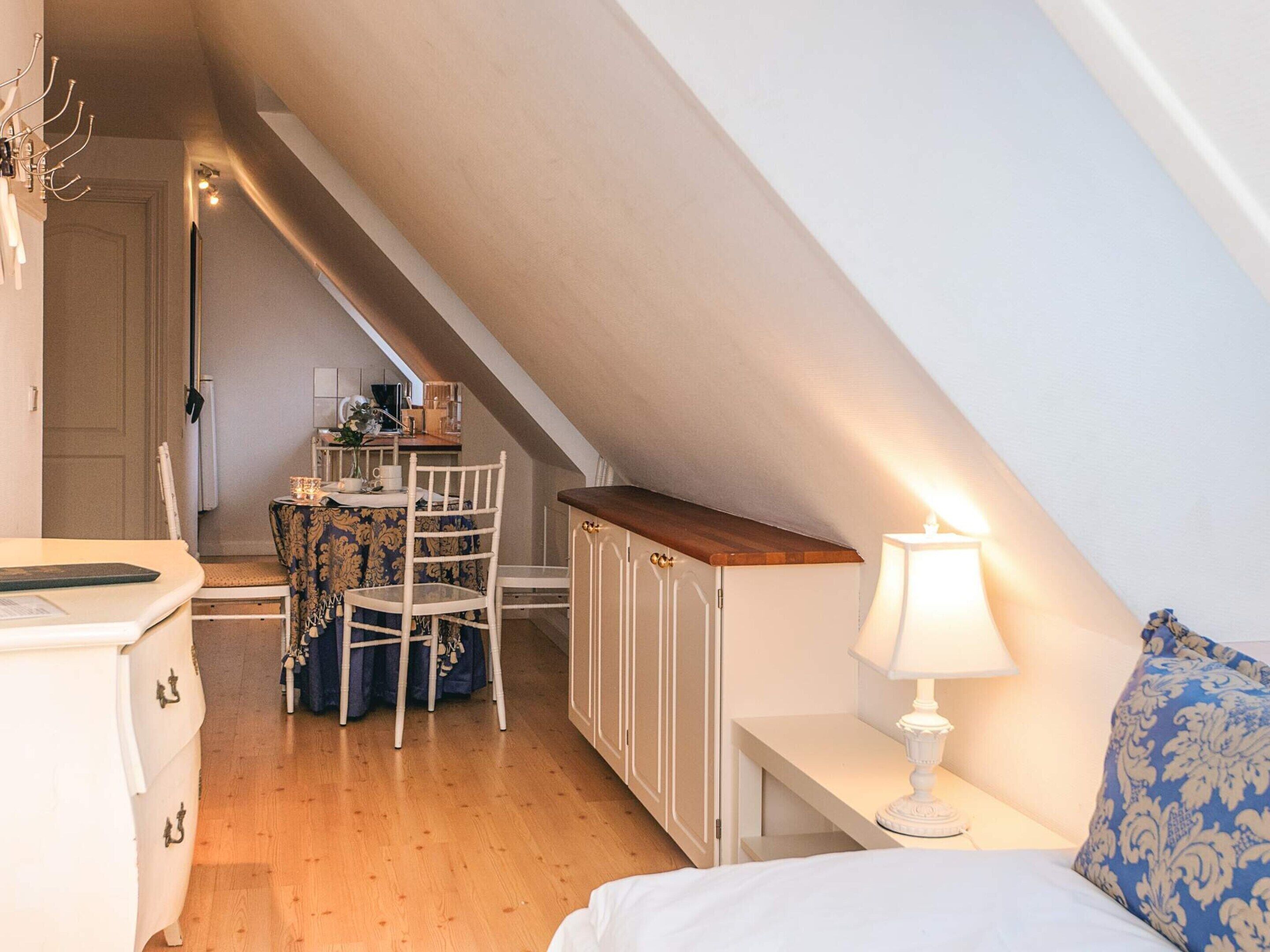 Un dormitorio acogedor en el ático, con techo inclinado, un escritorio de madera, una silla, una cama con un cojín estampado en azul y una mesita de noche con una lámpara.