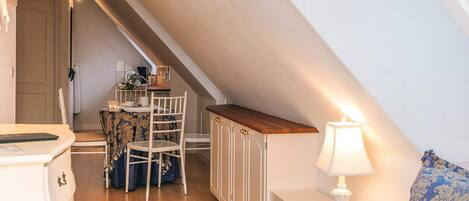 Une chambre avec un plafond en pente, un lit, une table de chevet avec une lampe, une chaise et une petite table sur laquelle est posé un vase.