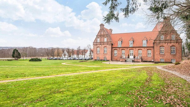 Een groot, historisch bakstenen gebouw met veel ramen en een opvallende toren, omringd door een verzorgd gazon en een parkeerplaats.