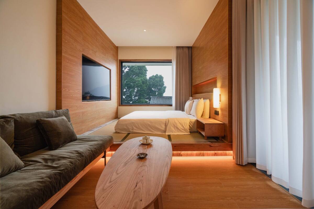 Una habitación de hotel moderna con un sofá, una cama, una mesa de centro de madera y un ventanal con vistas a árboles.