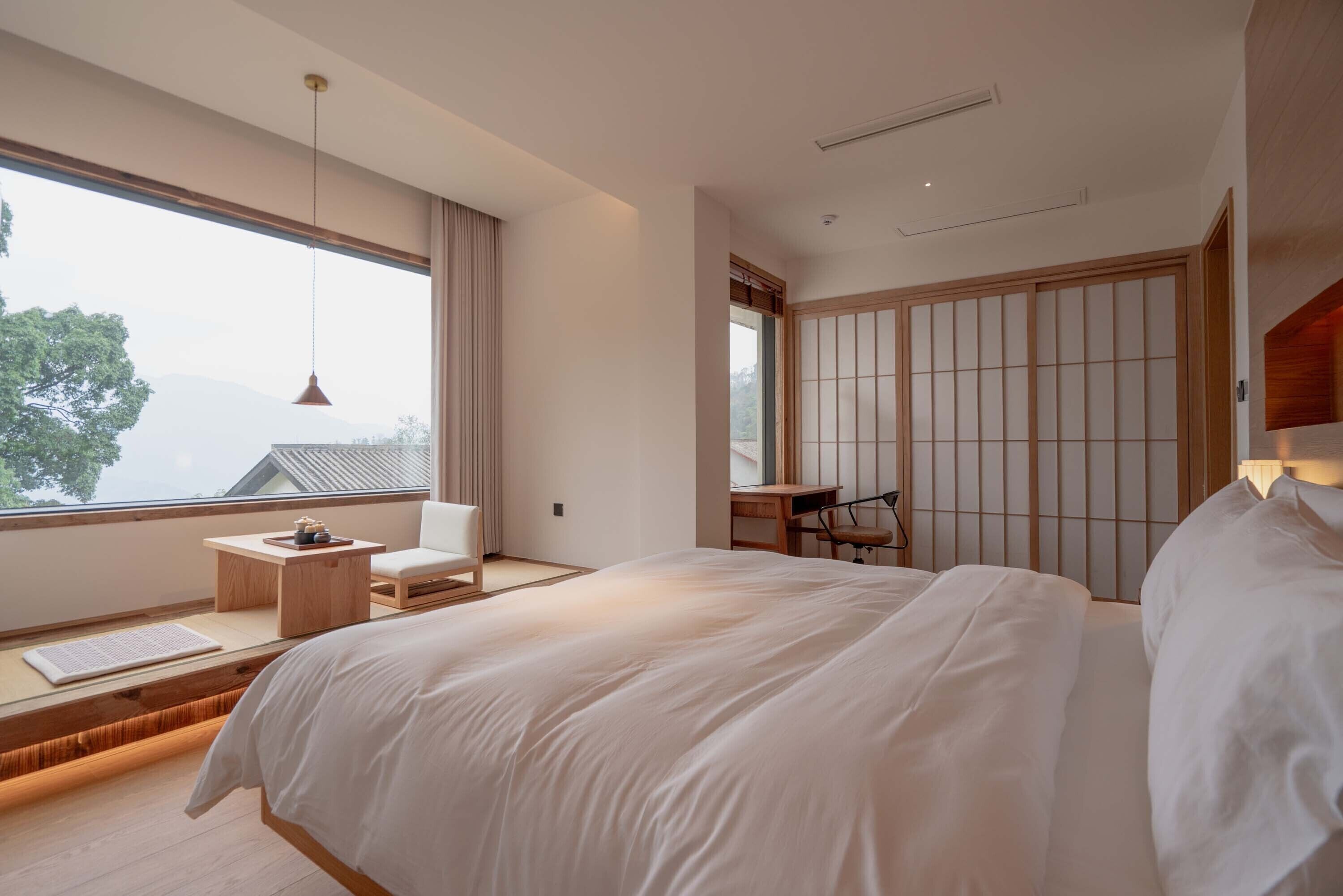 A modern bedroom with a large bed, a desk, and a chair. There is a window with a view of a mountain.