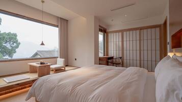 A modern bedroom with a large bed, a desk, and a chair. There is a window with a view of a mountain.
