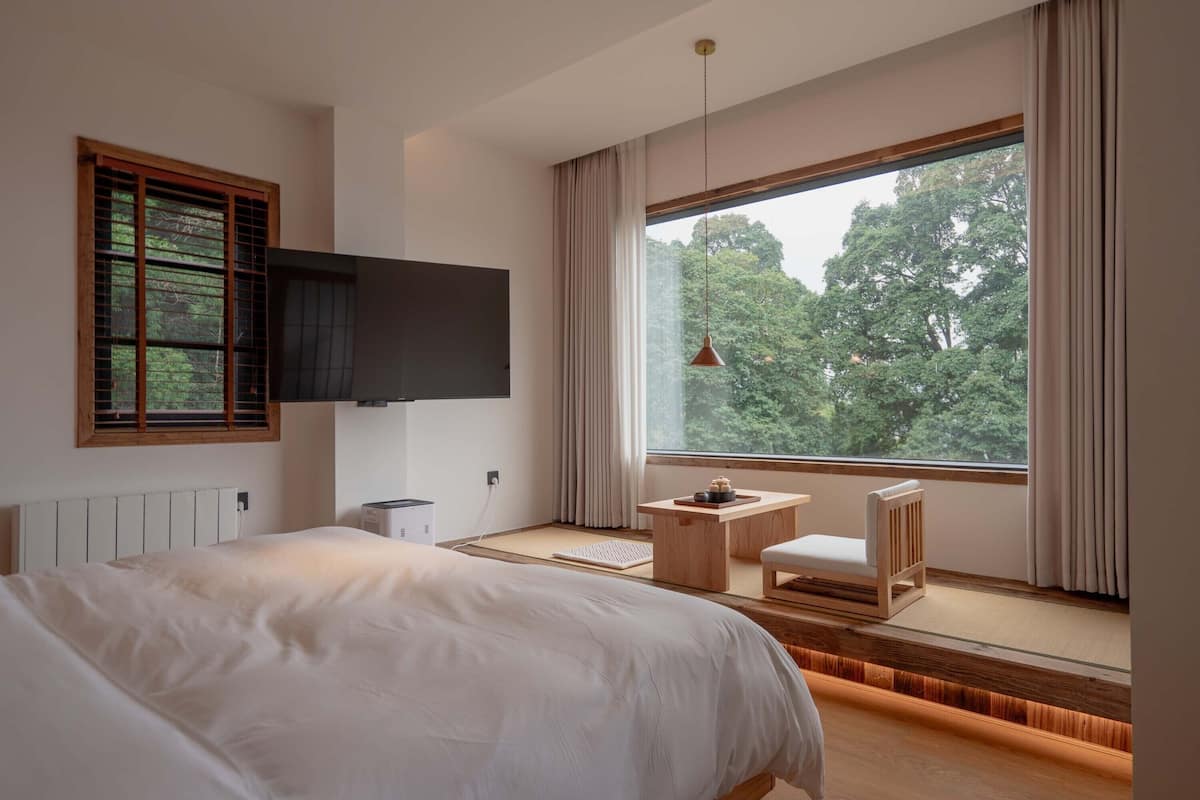 Un dormitorio con una cama grande, un televisor montado en la pared, una mesita con una silla y una ventana con vistas a árboles.