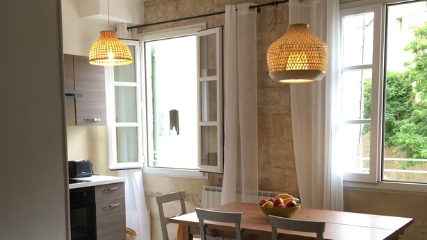 Ein Essbereich mit einem Holztisch, Korbstühlen und einer Obstschale. Zwei Hängelampen und ein Fenster mit Vorhängen sind vorhanden.