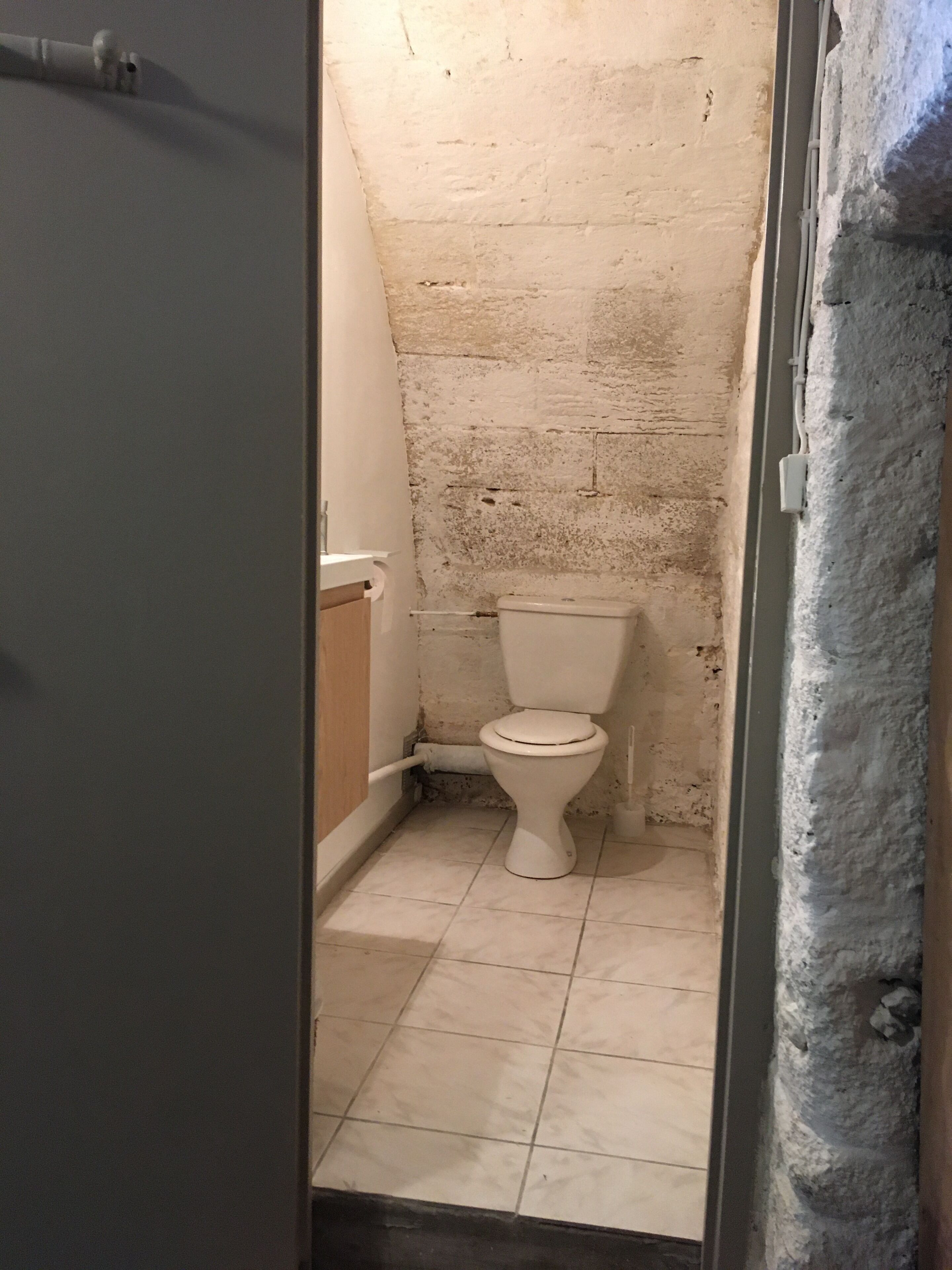 A small, narrow bathroom with a white toilet, tiled floor, and a rough-textured wall.