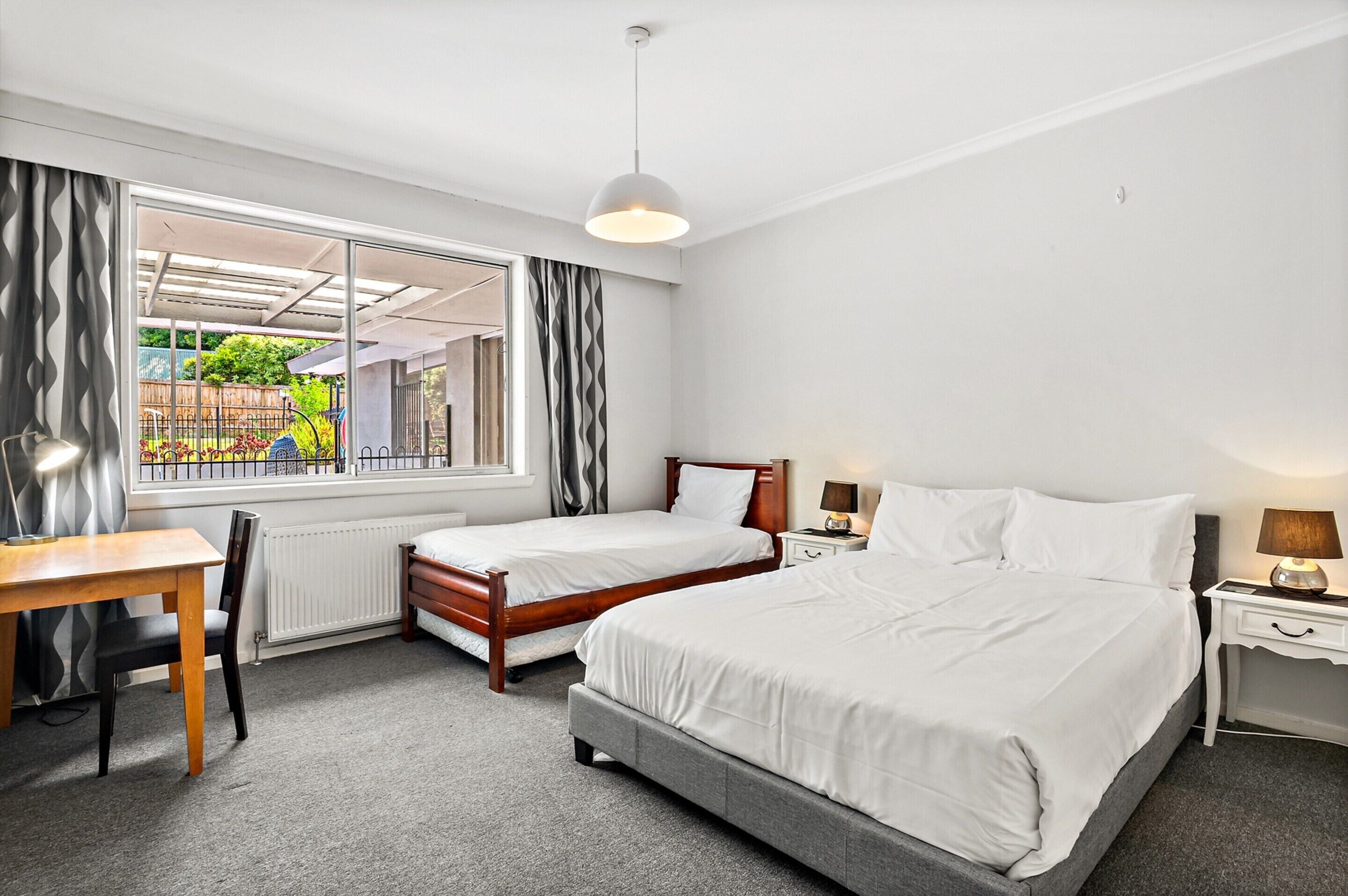 A hotel room with two single beds, a wooden desk, a chair, a bedside table with a lamp, and a view of a garden through a large window.