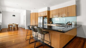 A modern kitchen with wooden cabinets, a central island with bar stools, and a dining area with a wooden table and chairs. - Spacious 5-bed Home With Pool in Glen Iris (Melbourne)