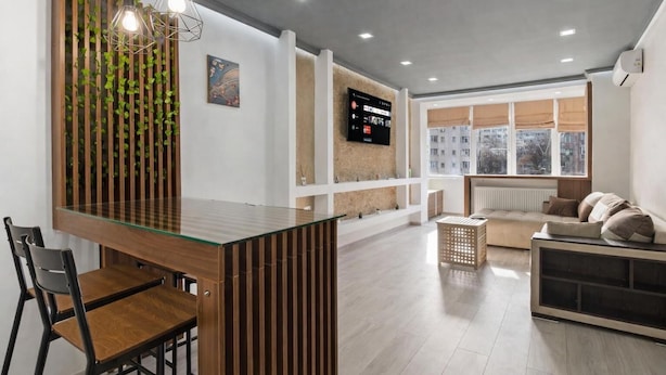 A modern living room with a glass dining table, wooden chairs, a sofa, and a TV mounted on the wall. - Apartament Lucas București  (Bucharest)