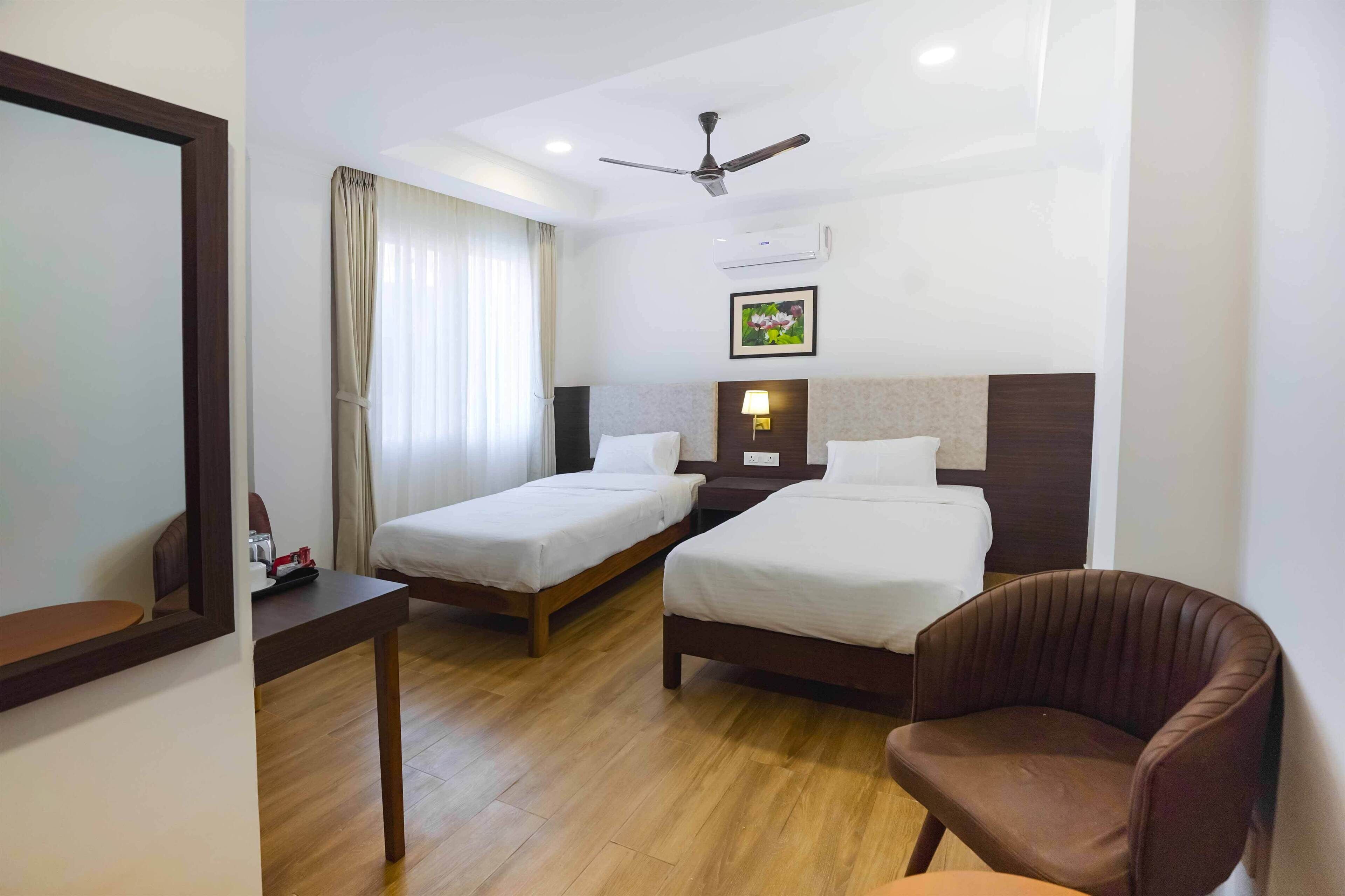 A hotel room with two single beds, a wooden desk, a brown leather chair, a ceiling fan, and an air conditioning unit.