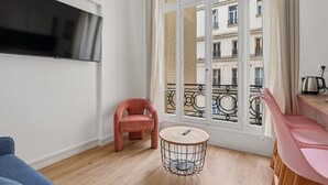 Una sala de estar moderna con televisor, una butaca rosa, una mesa de centro redonda y un balcón con vistas a edificios.