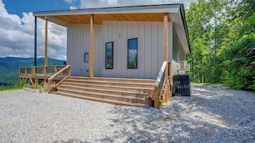 A two-story wooden cabin with a covered porch, a gravel driveway, and a mountain backdrop.
