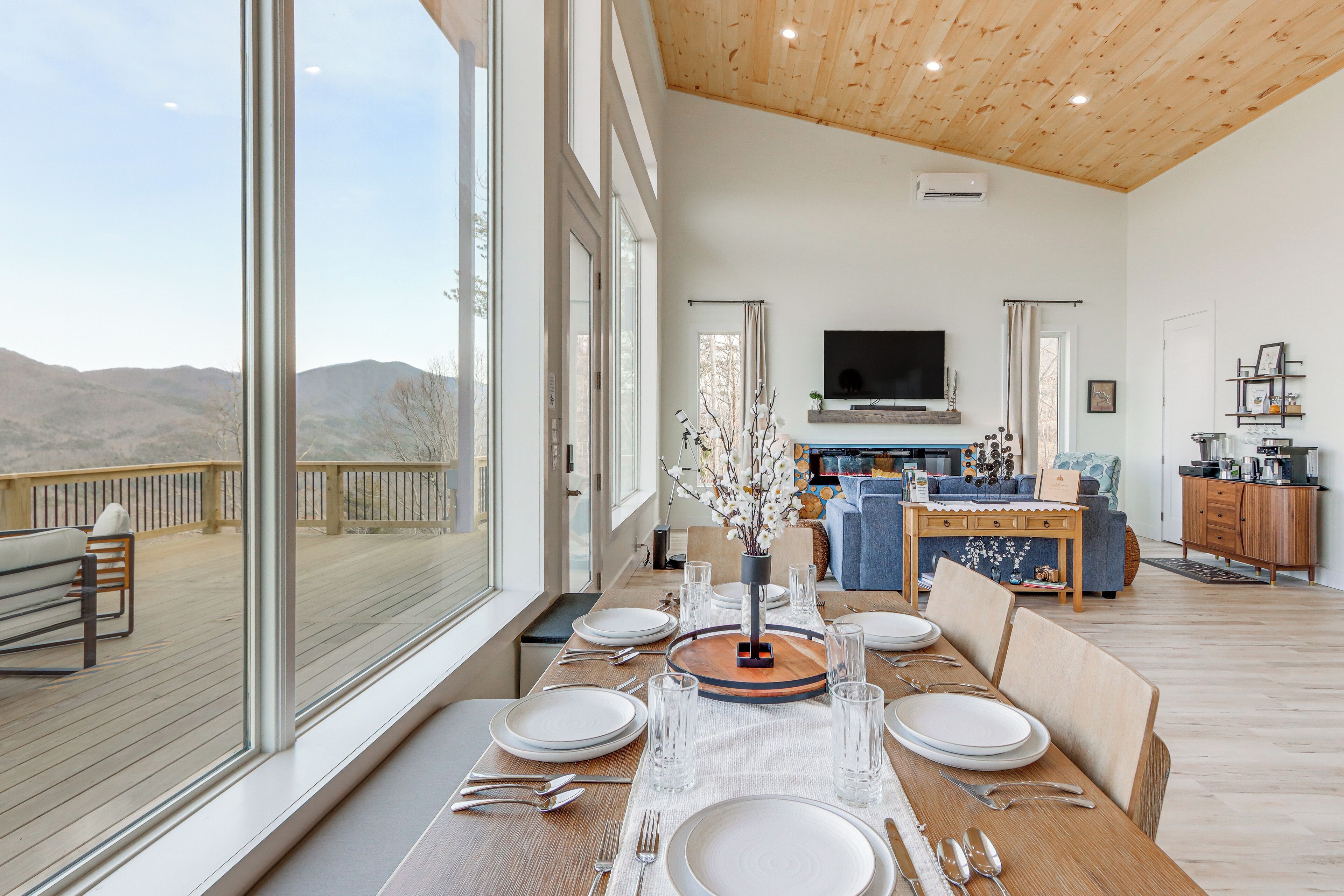 Un comedor con una mesa grande dispuesta para seis comensales, un televisor montado en la pared y vistas a las montañas a través de amplios ventanales.