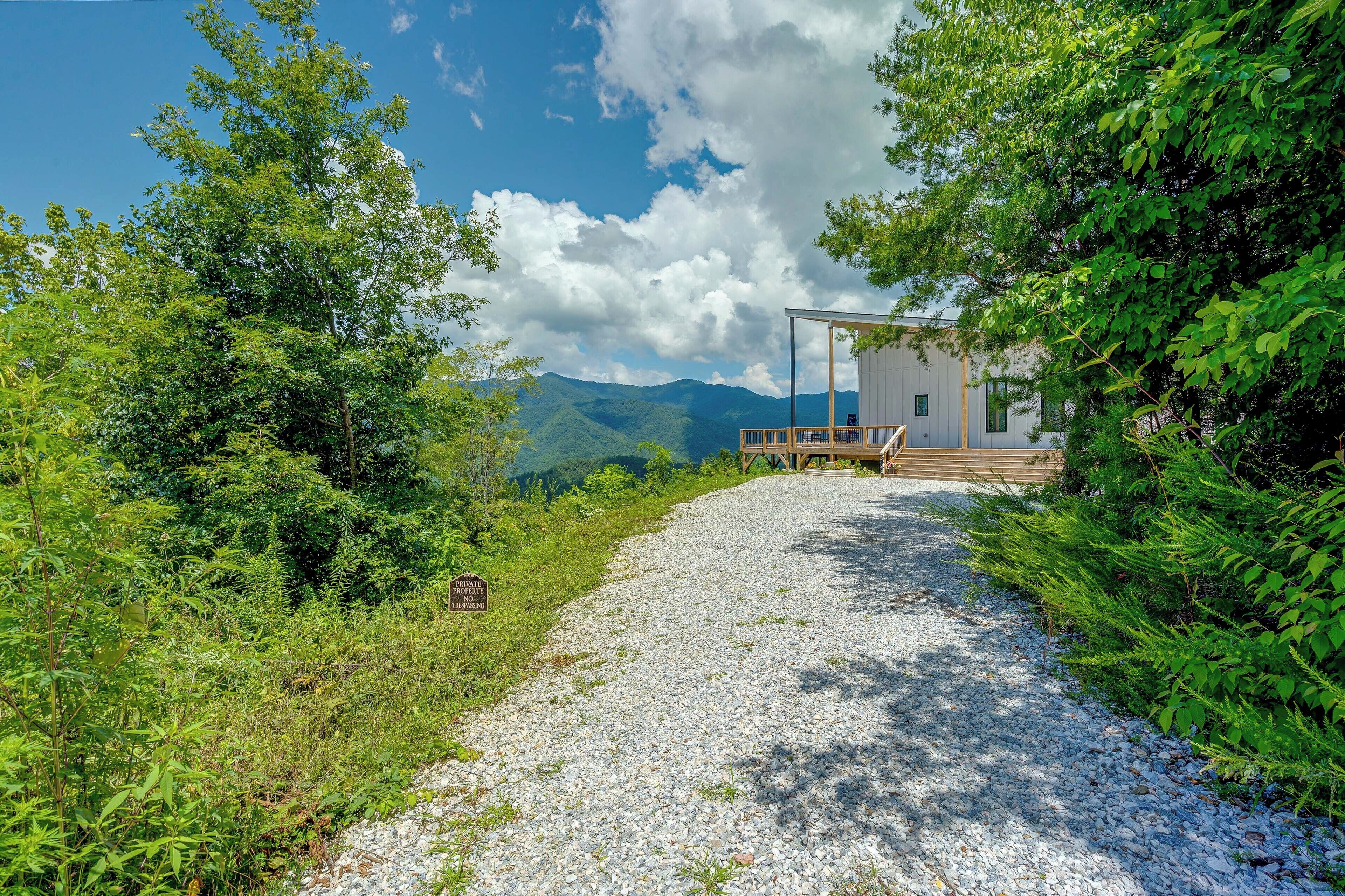 A gravel path leading to a wooden deck with a modern building, surrounded by lush greenery and mountains in the background.