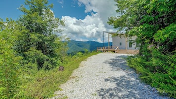 A gravel path leading to a wooden deck with a modern building, surrounded by lush greenery and mountains in the background.