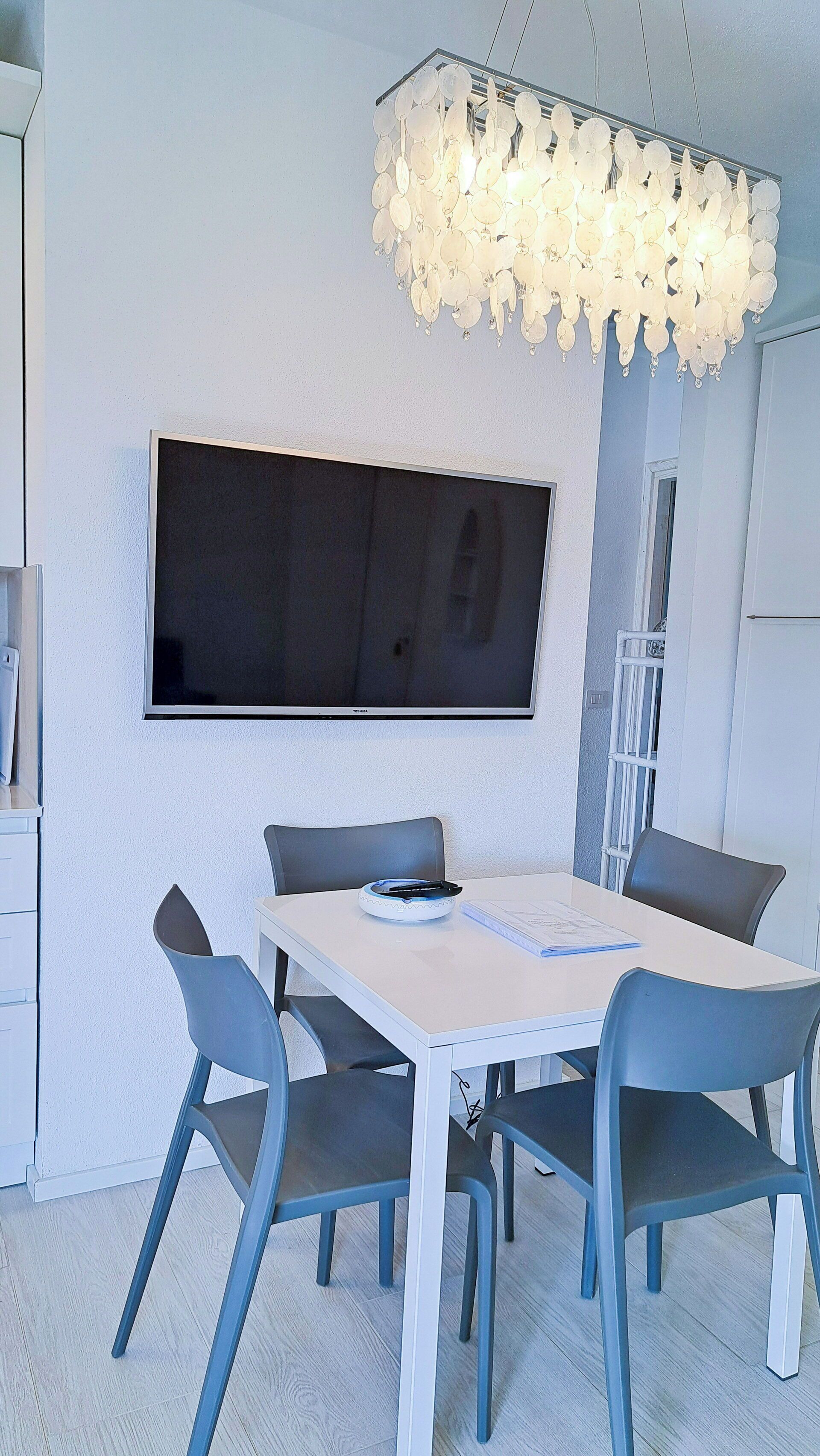 Un espace repas moderne avec une table blanche, quatre chaises grises, un téléviseur à écran plat fixé au mur et un lustre original.