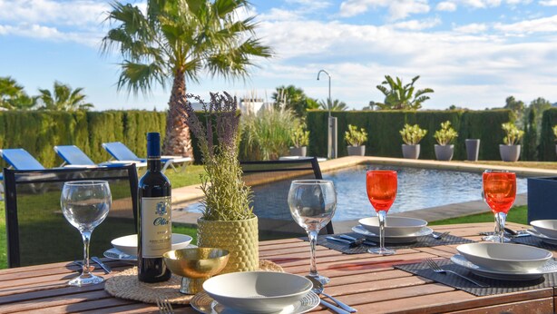 Mesa de comedor servida con copas de vino, platos y una botella, con vista a una piscina con palmeras y sillas de descanso.