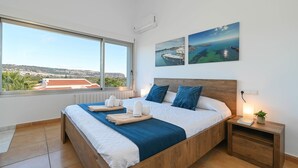 Un dormitorio moderno con una cama grande, una mesita de noche con lámpara y vista al mar a través de una ventana.
