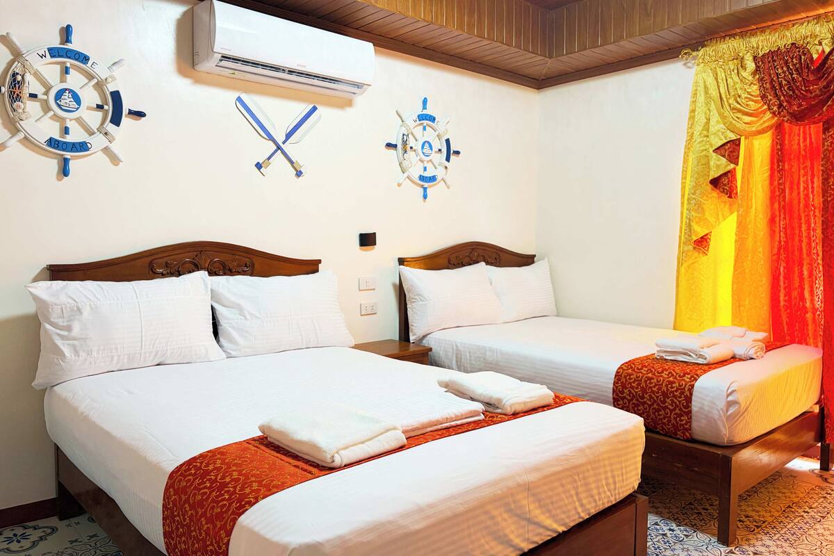 A hotel room with two beds, a ship's wheel decoration, an air conditioning unit, and a curtain with a golden and red pattern.