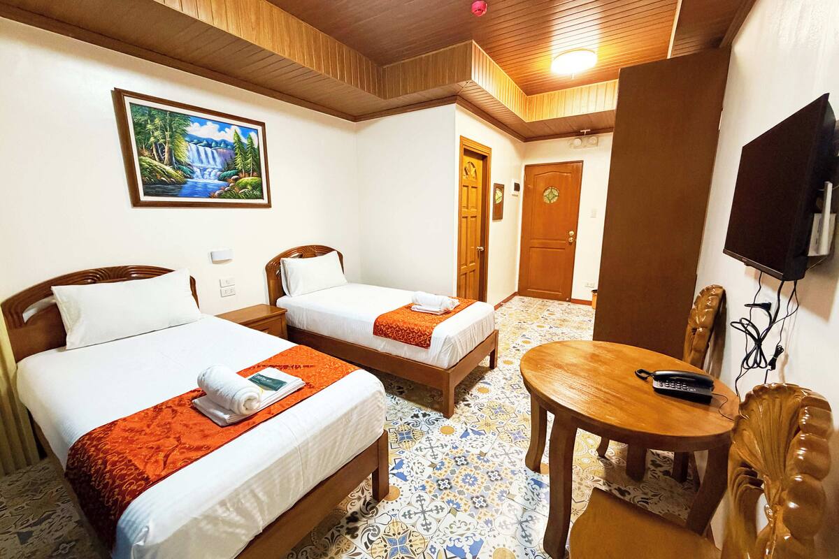 A hotel room with two beds, a wooden table, a TV, and a painting on the wall.
