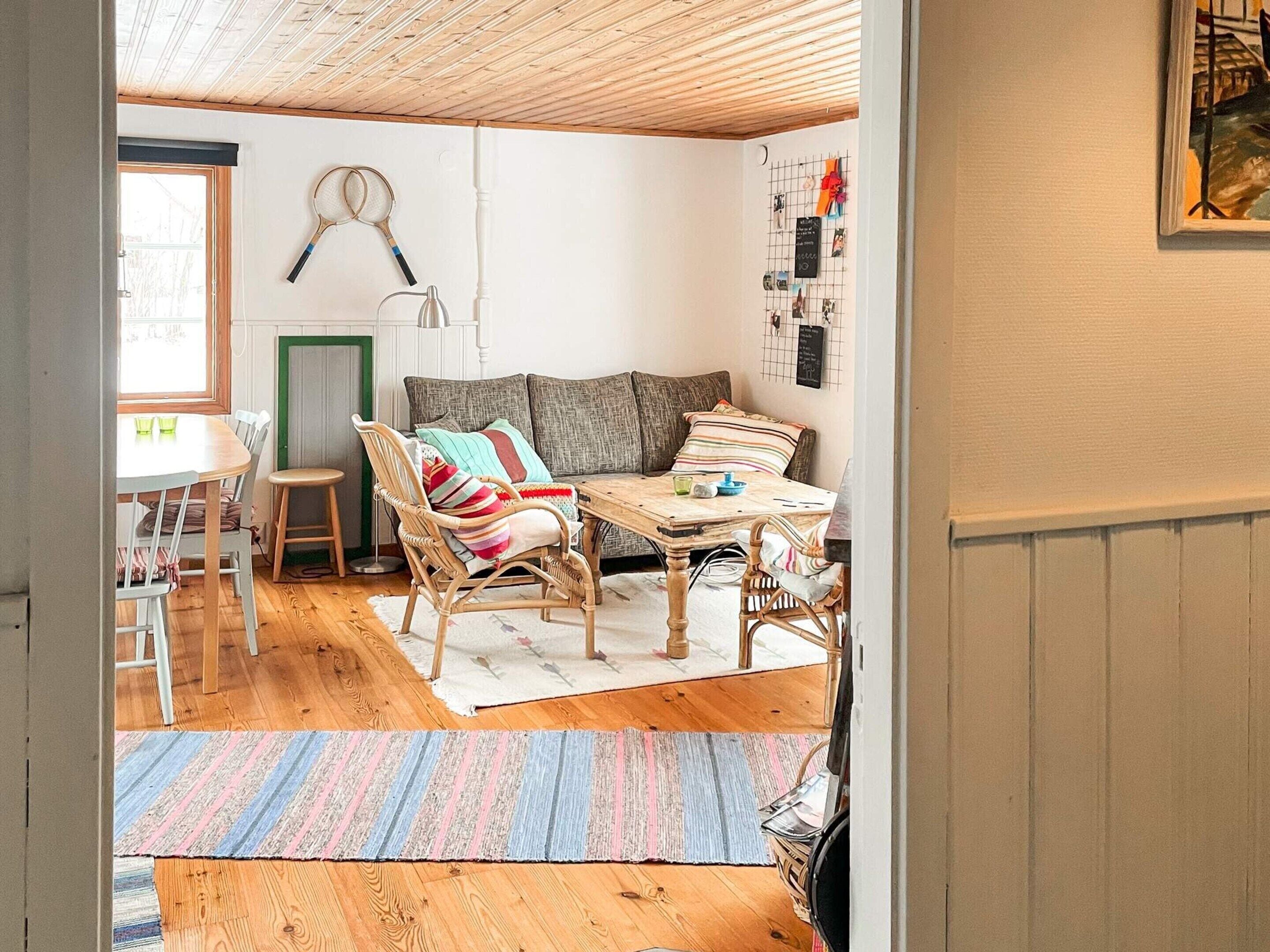 Um quarto aconchegante com teto de madeira, um sofá com almofadas coloridas, uma mesa de jantar com cadeiras e um suporte na parede com raquetes de badminton.