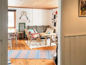 Um quarto aconchegante com teto de madeira, um sofá com almofadas coloridas, uma mesa de jantar com cadeiras e um suporte na parede com raquetes de badminton.