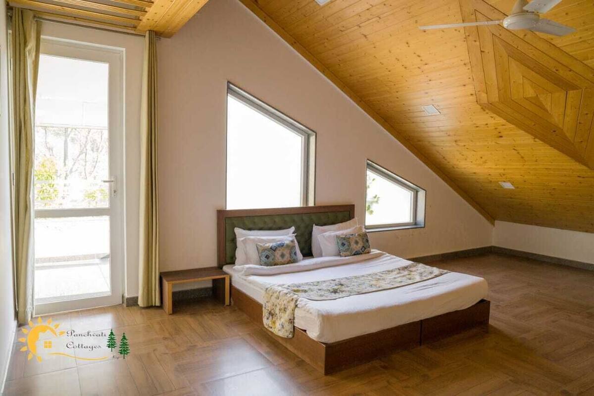A bedroom with a sloped ceiling, a bed with pillows, a bench, and large windows.