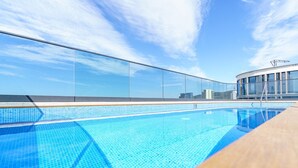 Une piscine sur le toit, avec une eau d’un bleu cristallin, entourée d’une barrière de verre et un bâtiment moderne en arrière-plan.