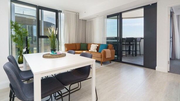 Un salon moderne avec une table blanche, des chaises noires, un canapé marron et une grande fenêtre.