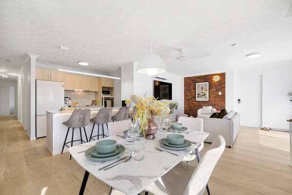 A modern kitchen with white cabinets, a central island with bar stools, and a dining area with a marble table set for four.