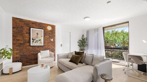 A modern living room with a brick accent wall, a white sofa, a round coffee table, and a balcony with a view of greenery.