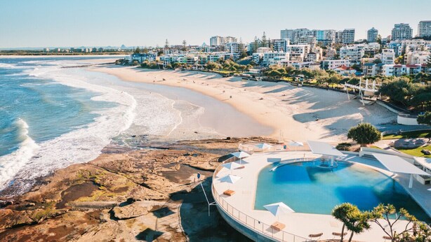 A coastal resort with a beach, a swimming pool, and a cityscape in the background.