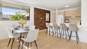 A modern kitchen with a brick accent wall, a dining area with a white table and grey chairs, and a view of the cityscape through large windows.