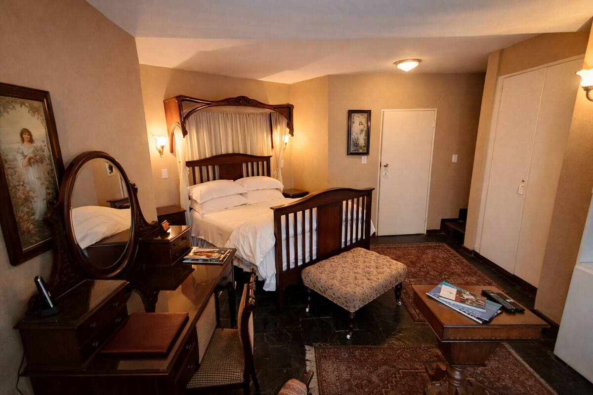 A hotel room with a four-poster bed, a bedside table with a lamp, a chair, a small desk with a chair, a rug, and a door.
