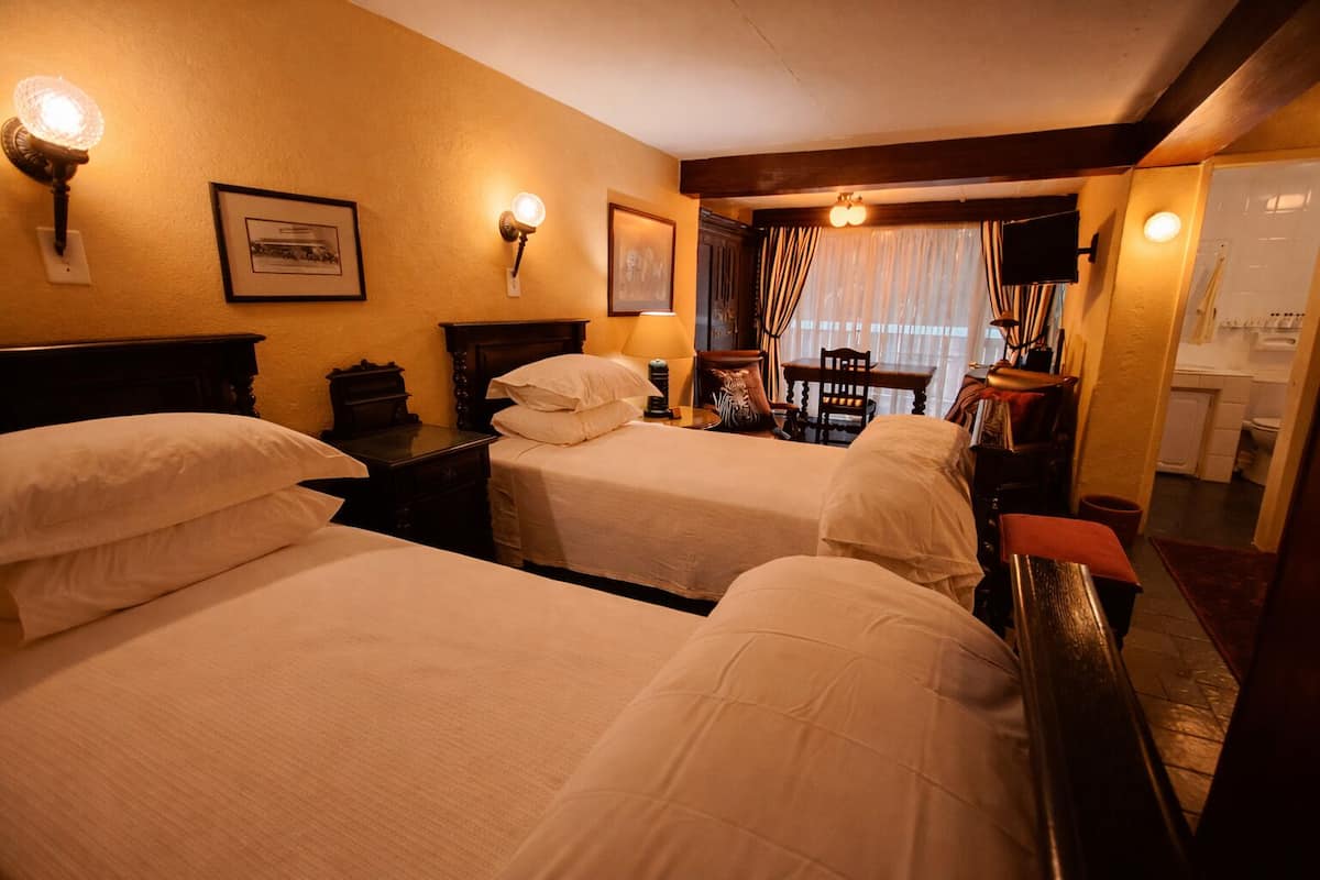 A hotel room with two beds, a desk, a chair, a small table, and a window with curtains.