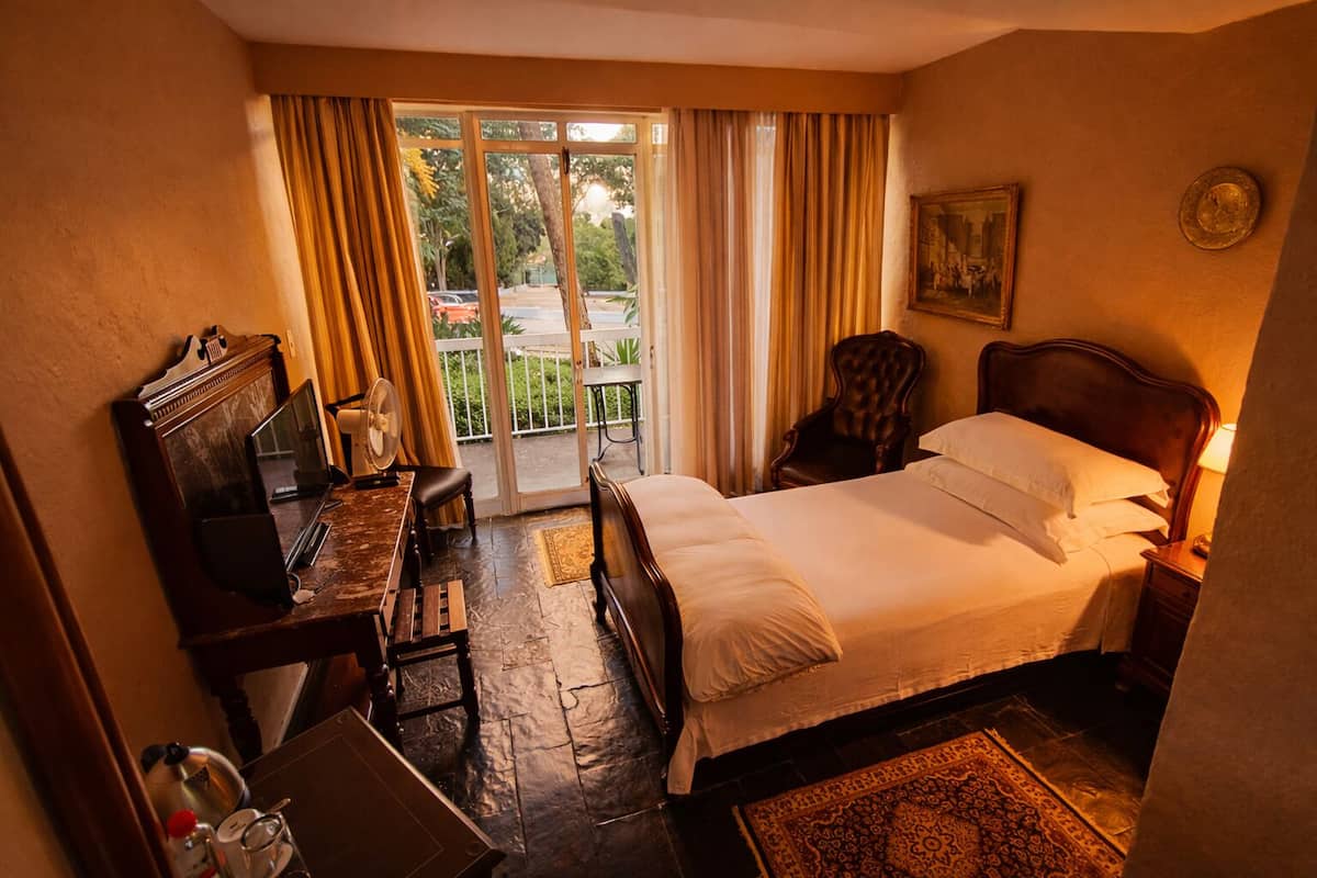 A hotel room with a bed, a desk with a mirror, a chair, a small table with a lamp, and a view of the outdoors through a sliding door.