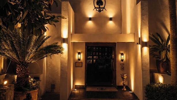 A building entrance with a lit emblem above the door, surrounded by potted plants and palm trees. - Whistletree Luxury Manor (Pretoria)