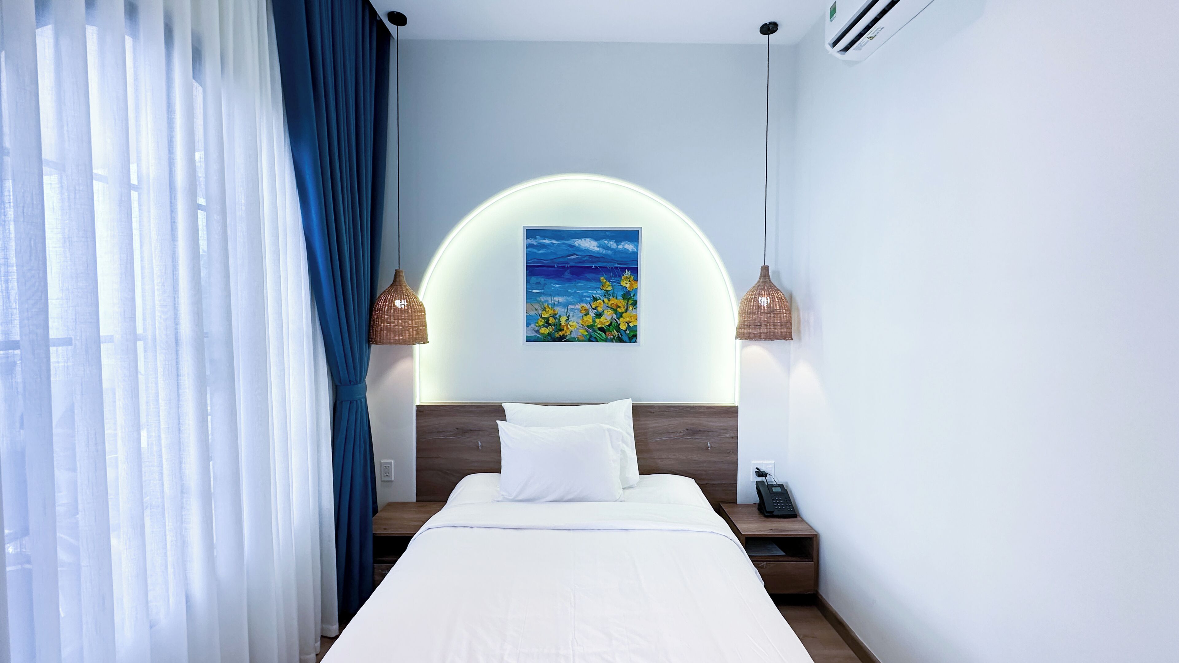 A hotel room with a bed, a painting, a phone on the nightstand, and a window with curtains.