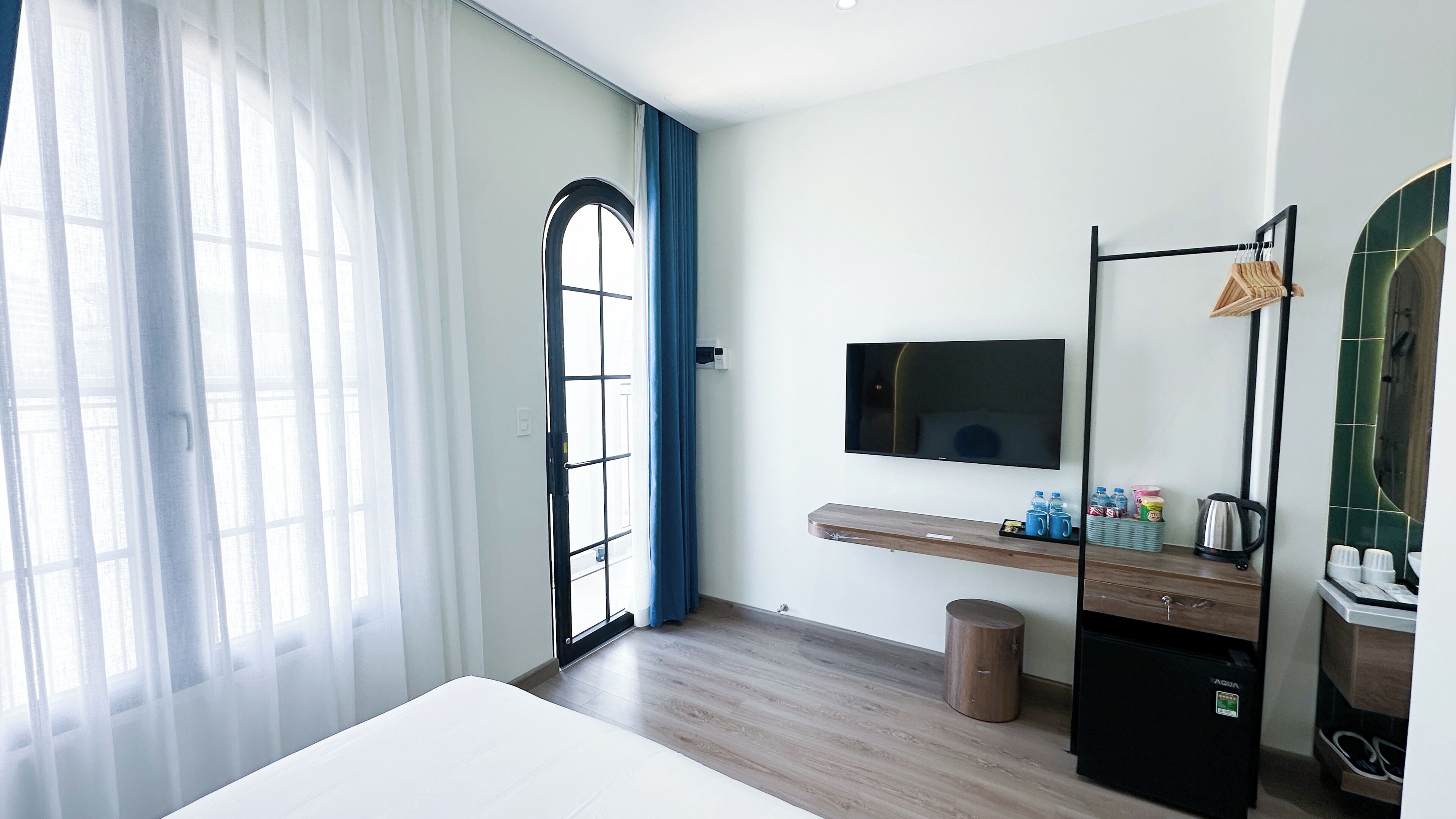 Quarto de hotel com cama, televisão na parede, penteadeira de madeira, lixeira pequena e janela com cortinas leves.
