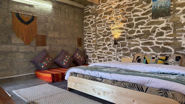 A rustic bedroom with a stone wall, wooden ceiling beams, a bed with patterned bedding, and colorful pillows. - Charming 3-bedroom B&B in serene Manali perfect for getaway (Naggar)