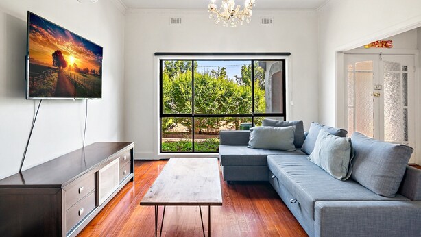 Een moderne woonkamer met een grijze bank, een flatscreen televisie, een houten salontafel en een groot raam met uitzicht op groen.