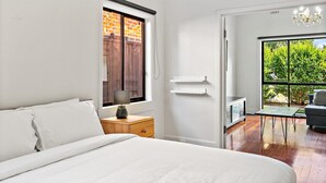 Een slaapkamer met een bed, nachtkastje, lamp, dressoir, een raam met uitzicht op een bakstenen muur, en een deur naar een balkon met uitzicht op groen.
