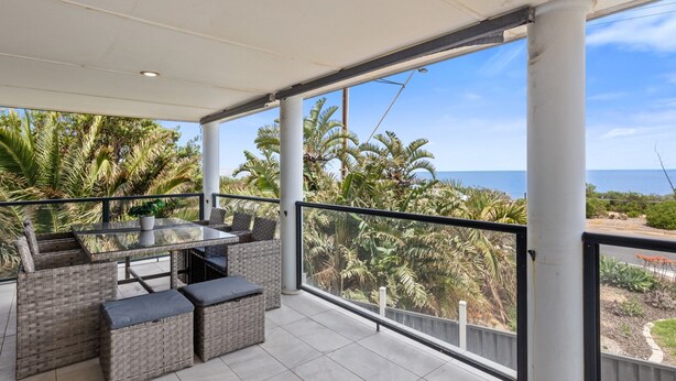 A covered outdoor balcony with wicker furniture, a glass table, and a view of palm trees and the sea. - Wine Region Coastal Retreat on The Esplanade (Adelaide)