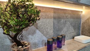Une salle de bain avec un petit bonsaï dans un vase en verre, un espace douche avec un porte-savon blanc, et quatre bouteilles violettes étiquetées « Shampoing » et « Gel douche ».