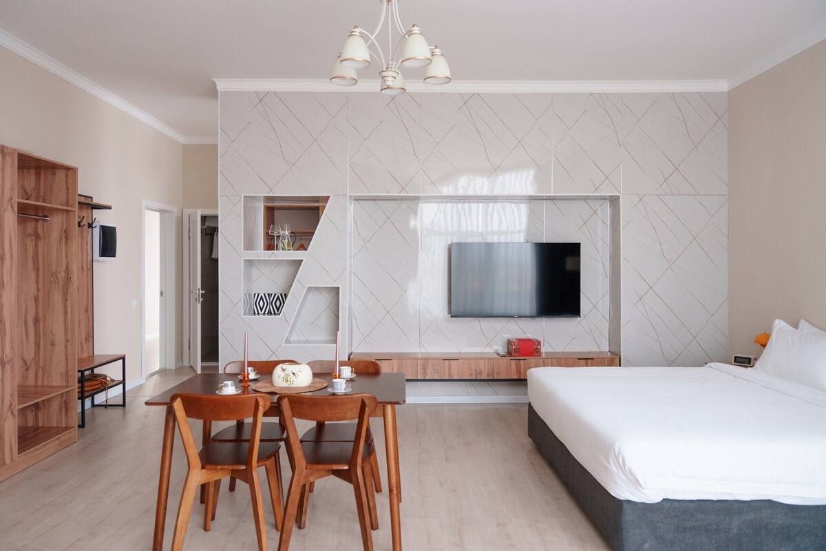 A modern hotel room with a large bed, a TV mounted on the wall, a dining table with chairs, and a wooden wardrobe.