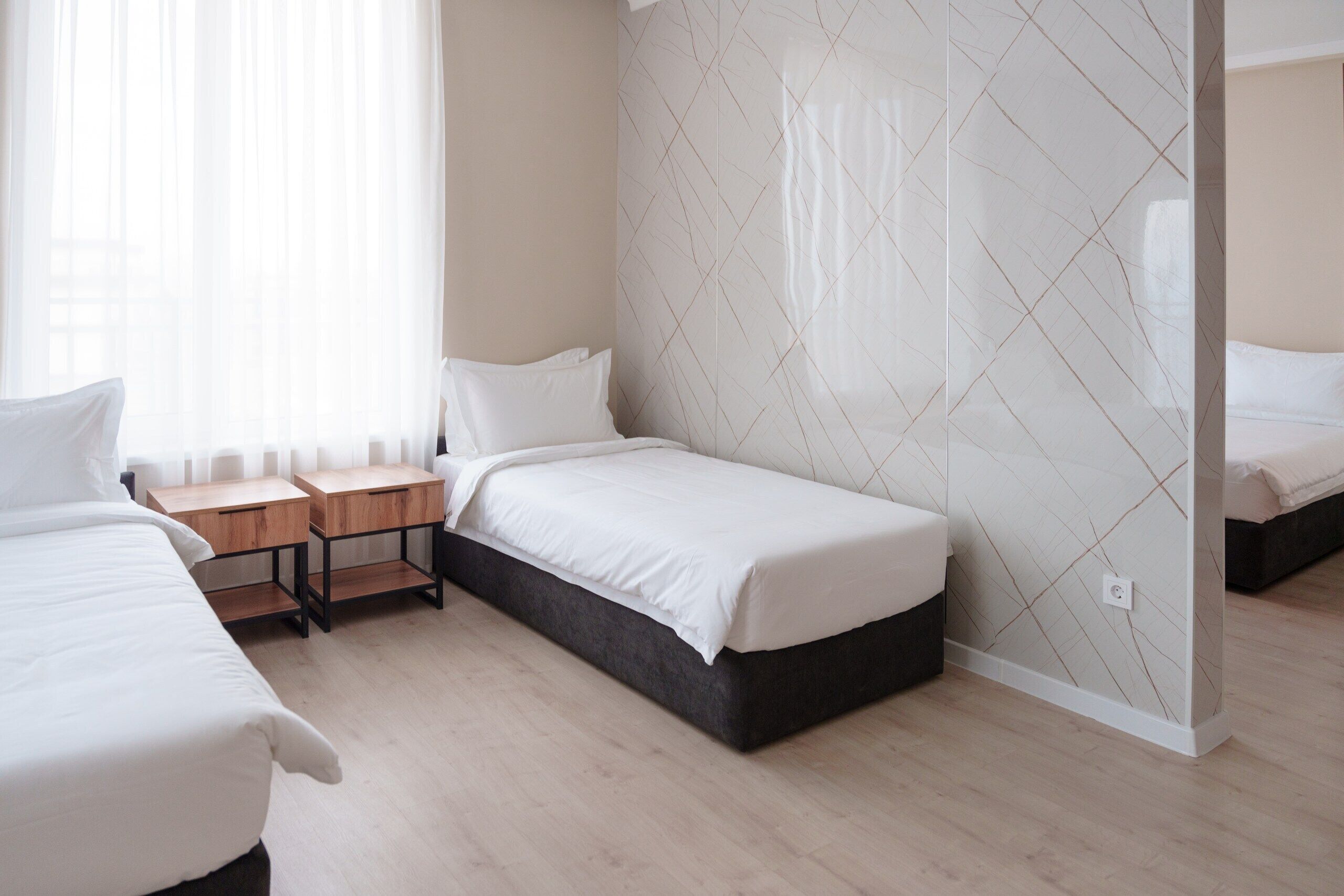 A modern hotel room with two beds, a wooden nightstand, and a large window with sheer curtains.